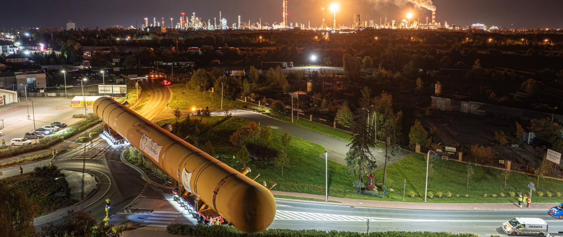 Transport ogromnego cylindrycznego elementu przemysłowego nocą, na tle oświetlonej rafinerii i instalacji przemysłowych.