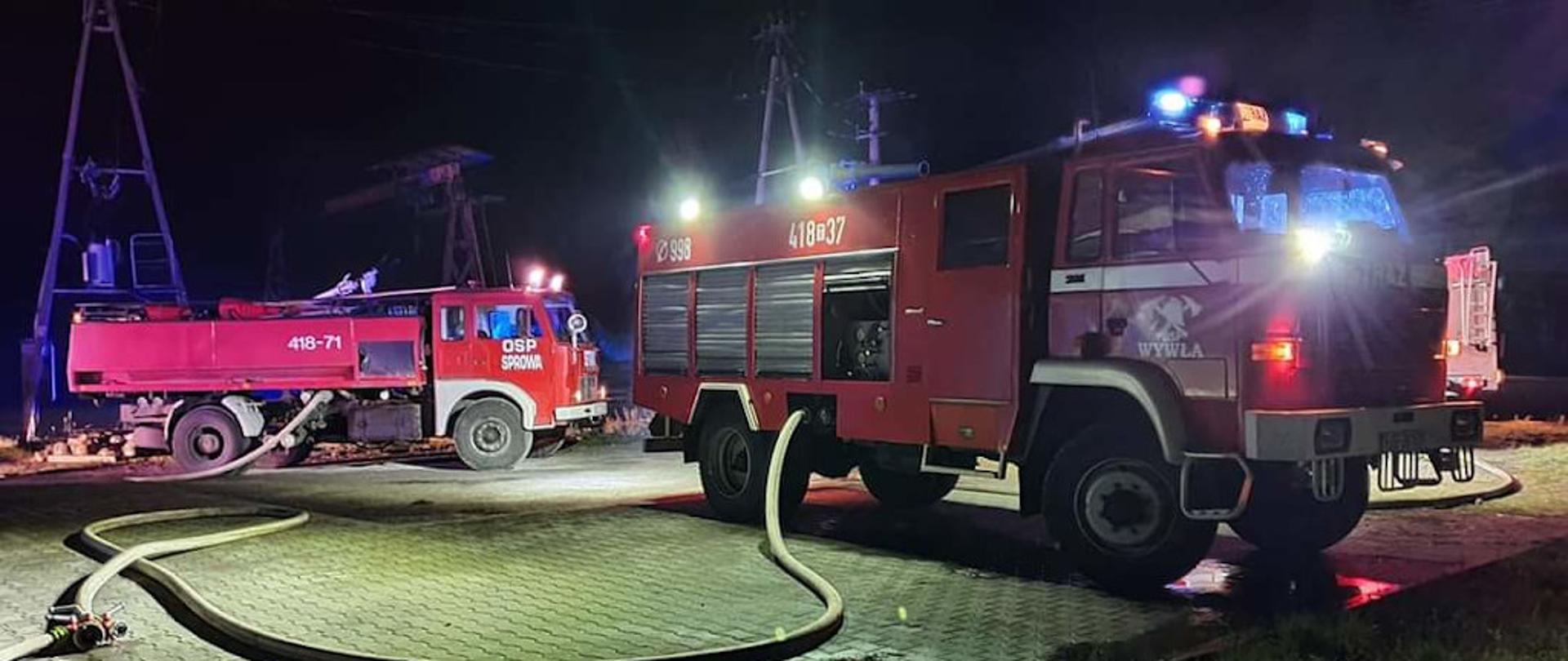 Na zdjęciu widoczne dwa pojazdy gaśnicze straży pożarnej. z włączonymi świetlnymi sygnałami przerywanymi (błyskowymi). Teren oświetlony ze względu na nocną porę działań ratowniczo- gaśniczych. Do samochodów podłączone są odcinki węży W-75.