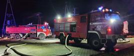 Na zdjęciu widoczne dwa pojazdy gaśnicze straży pożarnej. z włączonymi świetlnymi sygnałami przerywanymi (błyskowymi). Teren oświetlony ze względu na nocną porę działań ratowniczo- gaśniczych. Do samochodów podłączone są odcinki węży W-75.