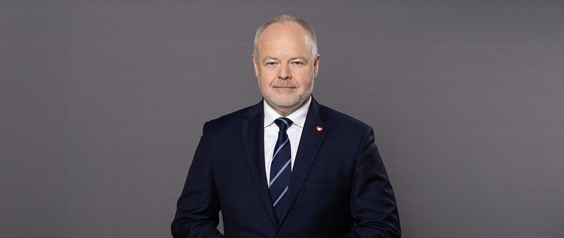 Wiceminister Marek Gzik stoi na tle ciemnoszarej ściany.