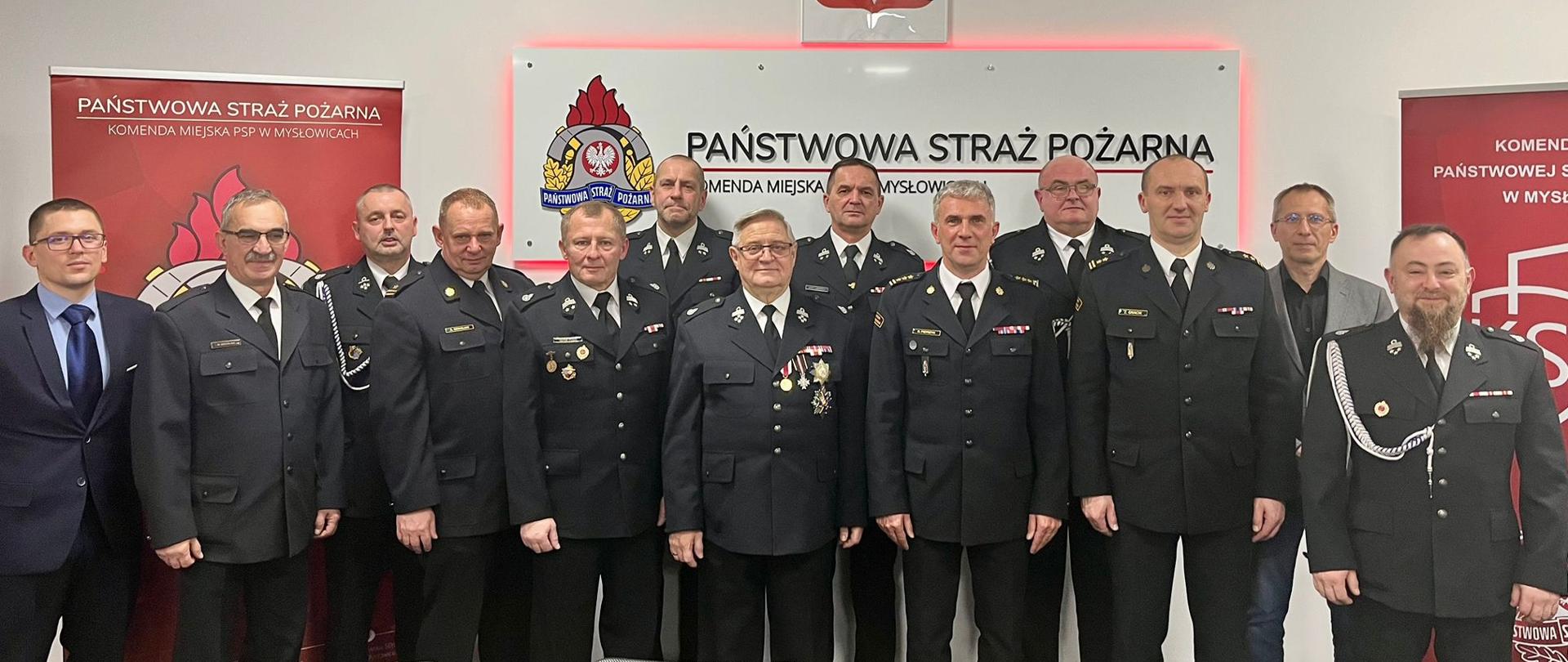Zdjęcie przedstawia grupę czternastu mężczyzn – oficerów Państwowej Straży Pożarnej (PSP) oraz przedstawicieli Ochotniczych Straży Pożarnych (OSP) – zgromadzonych w sali konferencyjnej Komendy Miejskiej Państwowej Straży Pożarnej w Mysłowicach.
Kontekst Wydarzenia:
* Spotkanie miało miejsce 11 grudnia 2025 roku w Komendzie Miejskiej Państwowej Straży Pożarnej w Mysłowicach.
* Było to spotkanie z Prezesami i Naczelnikami jednostek Ochotniczych Straży Pożarnych (OSP), które działają w ramach Krajowego Systemu Ratowniczo-Gaśniczego (KSRG).
* Wydarzenie zostało zorganizowane w związku z obchodami 30-lecia powstania KSRG, który jest fundamentalny dla krajowego systemu bezpieczeństwa i współpracy służb ratowniczych.
Uczestnicy i Ubiór:
* Większość mężczyzn ubrana jest w galowe mundury strażackie z dystynkcjami, medalami i odznaczeniami, co jest typowe dla uroczystych spotkań służb mundurowych.
* Wśród mężczyzn są zarówno funkcjonariusze PSP, jak i Prezesi/Naczelnicy jednostek OSP, co odzwierciedla cel spotkania: integrację i współpracę obu formacji.
* Trzech mężczyzn (jeden w garniturze po lewej i dwóch w odzieży cywilnej po prawej) stoi obok mundurowych.
Tło:
* Centralnym elementem tła jest biała tablica z logo i napisem „PAŃSTWOWA STRAŻ POŻARNA” oraz informacją o Komendzie Miejskiej.
* Powyżej widoczny jest Orzeł Biały, godło Rzeczypospolitej Polskiej.
* Po bokach widoczne są czerwone banery z logo PSP, co potwierdza lokalizację jako Komenda Miejska PSP w Mysłowicach.
Całość jest pamiątkową fotografią z ważnego spotkania poświęconego współpracy PSP i OSP w ramach 30-lecia KSRG.