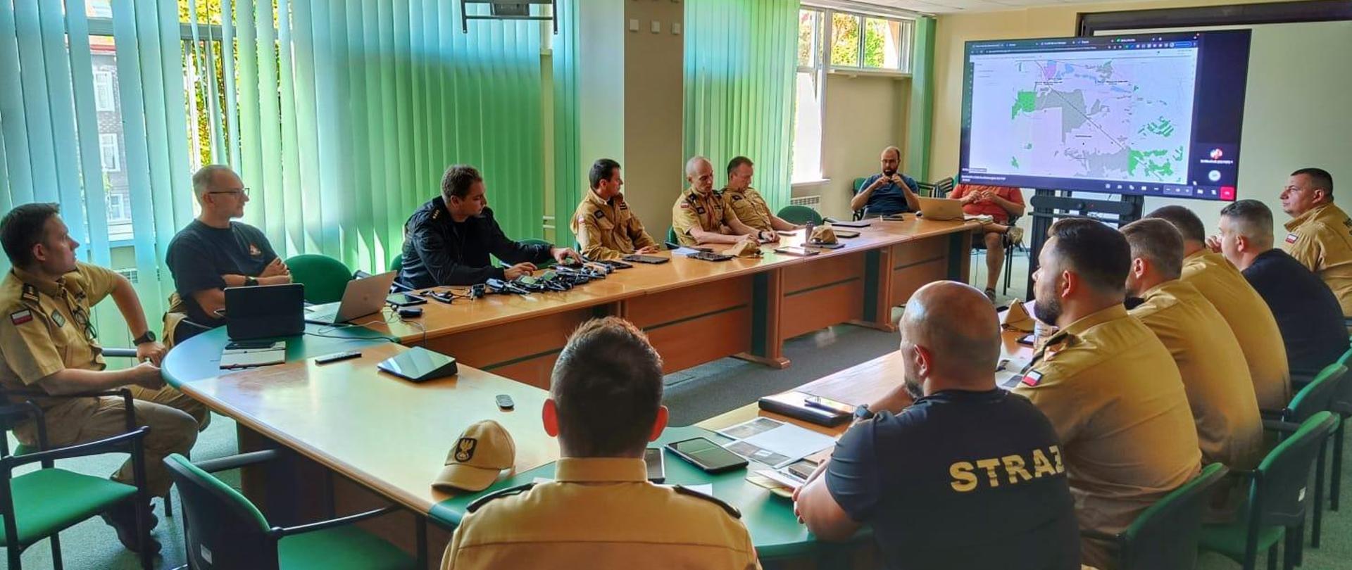 Grupa strażaków odbywa spotkanie w sali konferencyjnej. Na biurkach leżą telefony i radiotelefony. Pomiędzy stołami stoi telewizor wyświetlający prezentację.