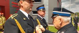 na zdjęciu plac przed budynkiem Komendy Powiatowej PSP w Brzezinach podczas uroczystej zbiórki z okazji Dania Strażaka, sekretarz zarządu Oddziału Wojewódzkiego ZOSP RP w Łodzi wręcza dwóm funkcjonariuszom komendy odznaczenie "Srebrny Medal za Zasługi dla Pożarnictwa""