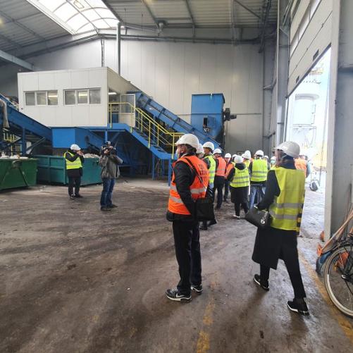 Osoby w kamizelkach odblaskowych i kaskach stoją w dużym, magazynowym pomieszczeniu.