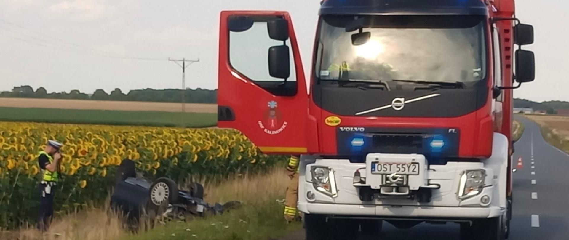 Na zdjęciu znajduje się wrak samochodu osobowego odwrócony kołami do góry, w tle widać pole słoneczników, wóz strażacki oraz policjanta.