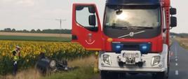Na zdjęciu znajduje się wrak samochodu osobowego odwrócony kołami do góry, w tle widać pole słoneczników, wóz strażacki oraz policjanta.