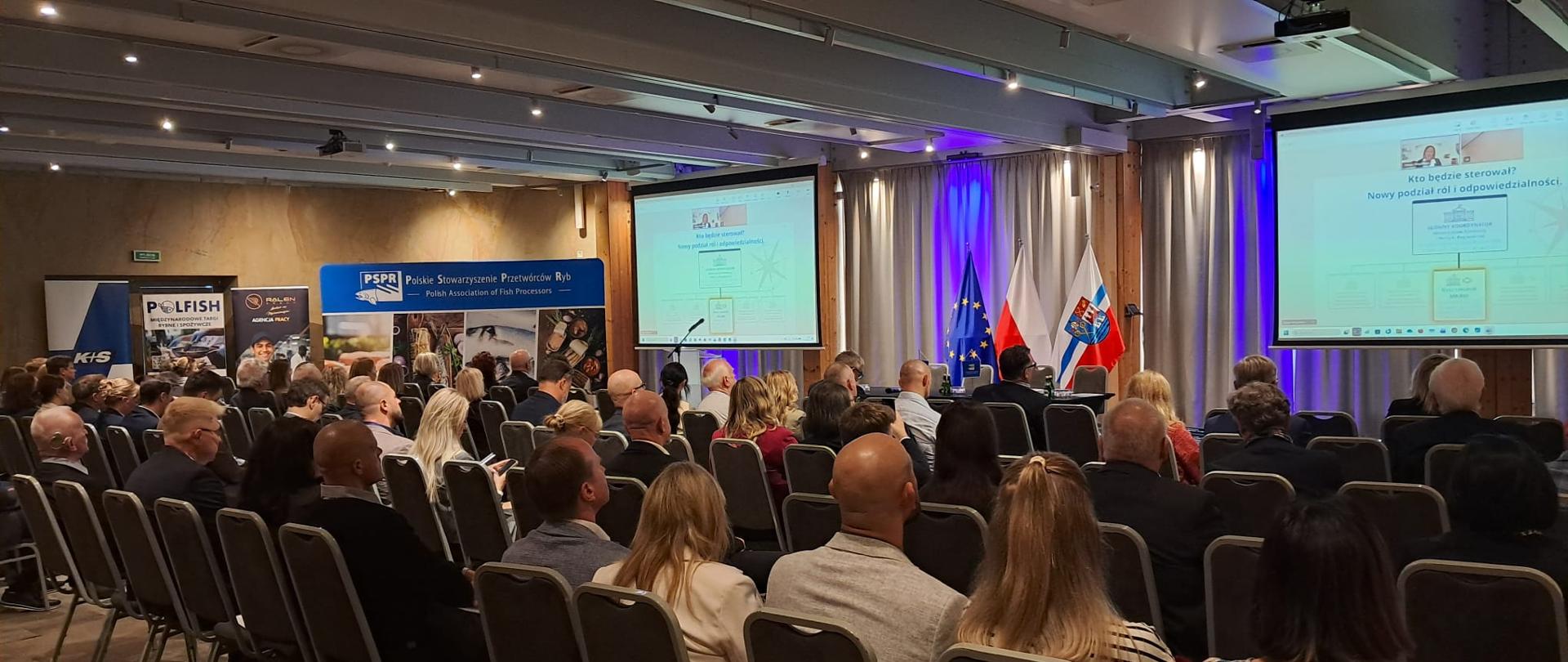 Sala konferencyjna wypełniona słuchaczami siedzącymi w rzędach, skierowanymi w stronę sceny. W tle nowoczesne tablice multimedialne wyświetlające prezentację biznesową oraz stojące banery promocyjne. Jasne oświetlenie, profesjonalna atmosfera wydarzenia szkoleniowego lub konferencji