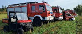 Samochody pożarnicze biorące udział w ćwiczeniach stoją na łące w miejscu zbiórki w oddali widać strażaków.