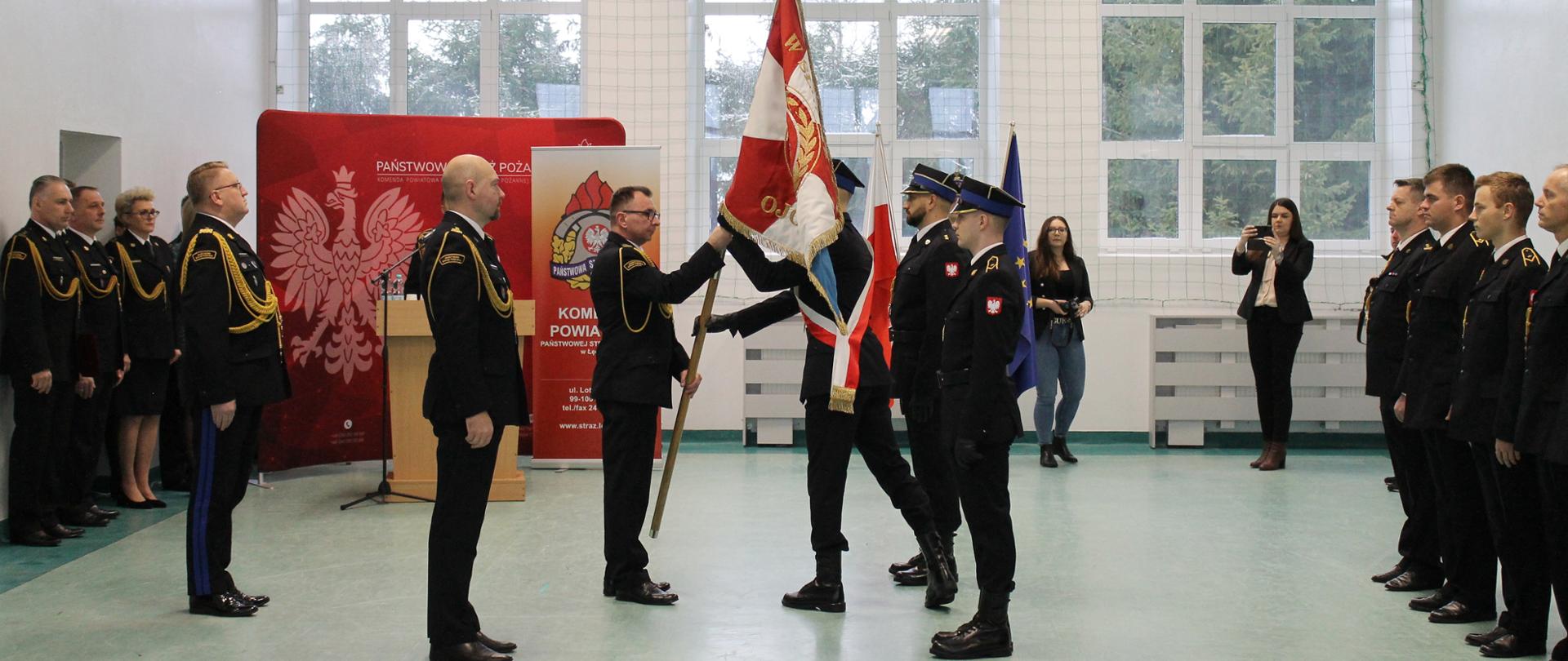 Na zdjęciu widać strażaków w mundurach oraz osoby cywilne stojące wewnątrz budynku. Sześciu strażaków stoi na środku a reszta pod ścianą. Trzech strażaków, w poczcie sztandarowym ma na głowie czapki. Jeden strażak przekazuje drugiemu poczet sztandarowy. W tle widać banery z grafiką i napisami strażackimi. W pomieszczeniu jest zapalone światło. Za oknem widać drzewa. Jest widno.