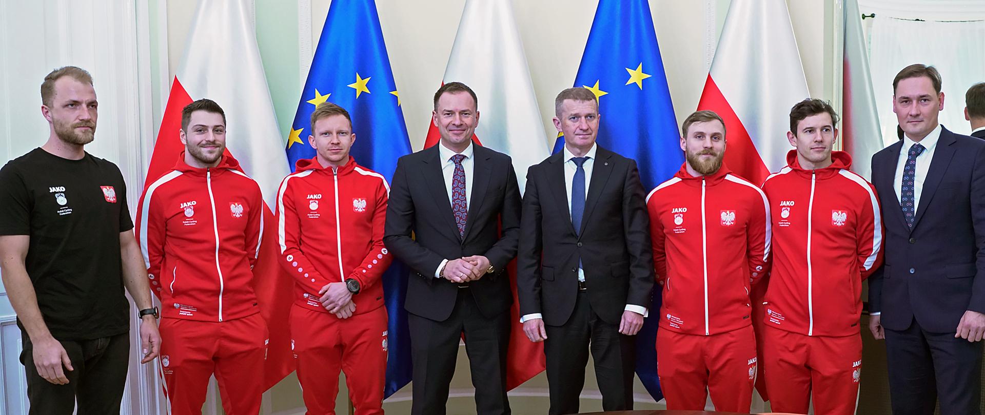 Spotkanie wiceministrów Piotra Borysa i Ireneusza Rasia z medalistami Mistrzostw Europy w Kolarstwie Torowym - ministrowie stoją na tle flag Polski i UE z medalistami, trenerem i przedstawicielem PZKol
