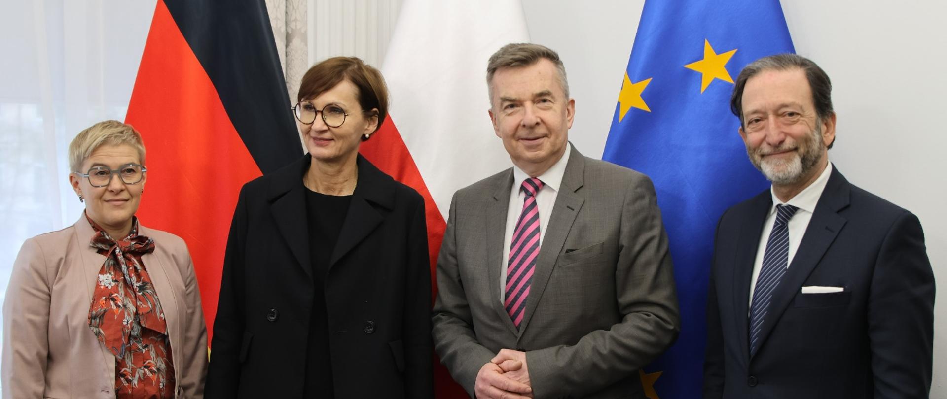 Minister Wieczorek i wiceminister Mrówczyńska stoją obok mężczyzny w czarnym garniturze i kobiety w czarnej marynarce, za nimi na tle ściany flagi Polski, Niemiec i UE.