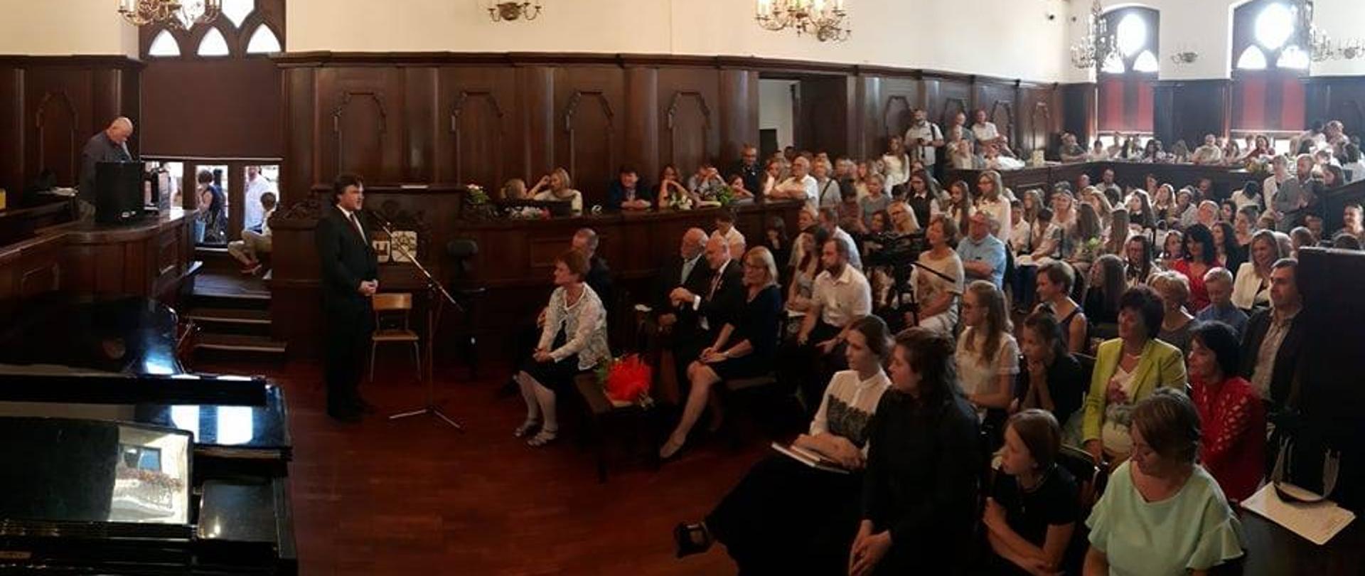 Zdjęcie uroczystości zakończenie roku szkolnego w sali Ratusza Miejskiego. Ściany ozdobione zabytkową dębową boazerią, sufit biały, zdobiony kasetonami ze złoconymi ornamentami i ozdobnymi żyrandolami. Po lewej stronie przemawiający do mikrofonu dyrektor, po prawej stronie zgromadzona publiczność. W lewym dolnym narożniku zdjęcia widoczny otwarty fortepian.
