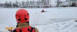 Strażak w czerwonym kombinezonie zanużony do połowy w przeręblu. Przypięty do liny asekuracyjnej. Drugi strażak pomaga mu się wydostać na sanie lodowe. Kolejny strażak przy linie asekuracyjnej. W tle linia brzegowa jeziora, drzewa oraz parking dla samochodów ciężarowych.