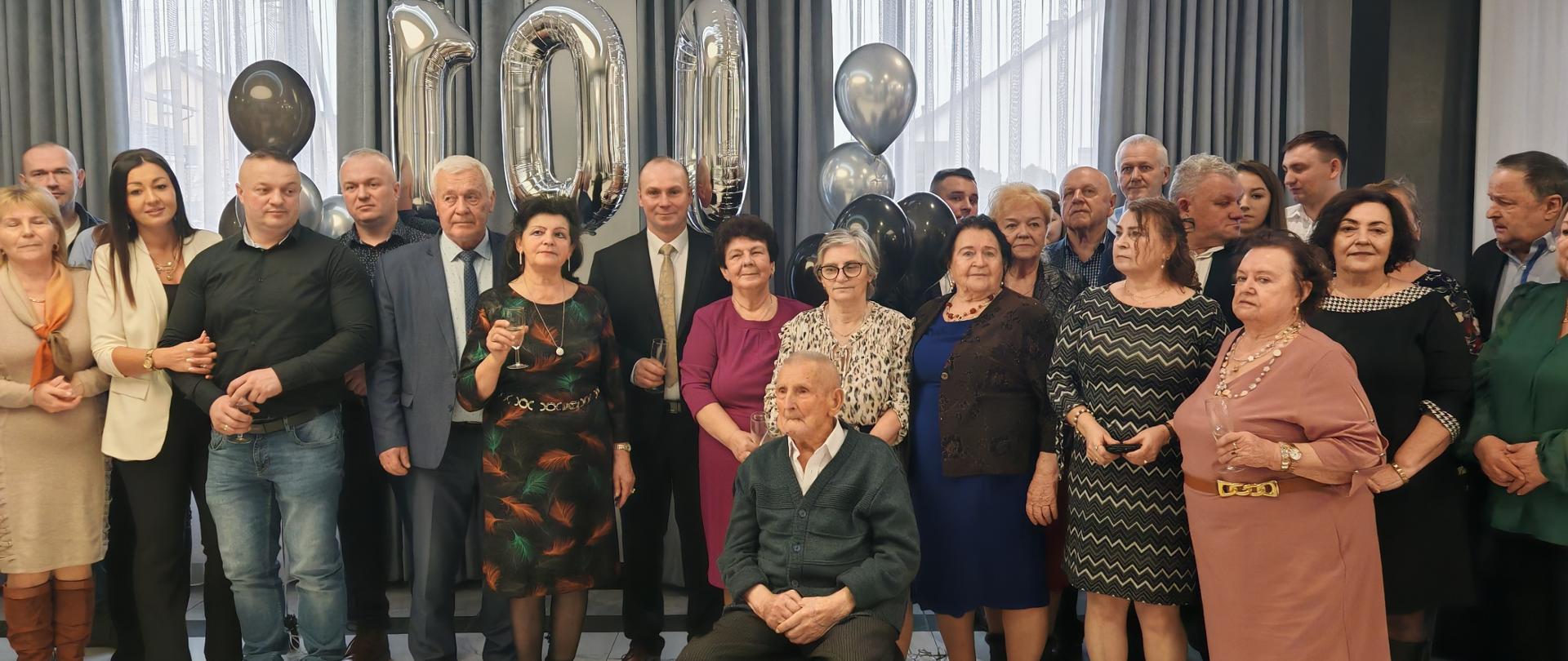 W jasnej sali stoi duża grupa dorosłych i seniorów, elegancko ubranych. Za nim wiszą srebrne balony tworzące liczbę ”100”. Na pierwszym planie siedzi starszy mężczyzna – jubilat. Wszyscy pozują blisko siebie, jak na uroczystej rodzinnej fotografii.