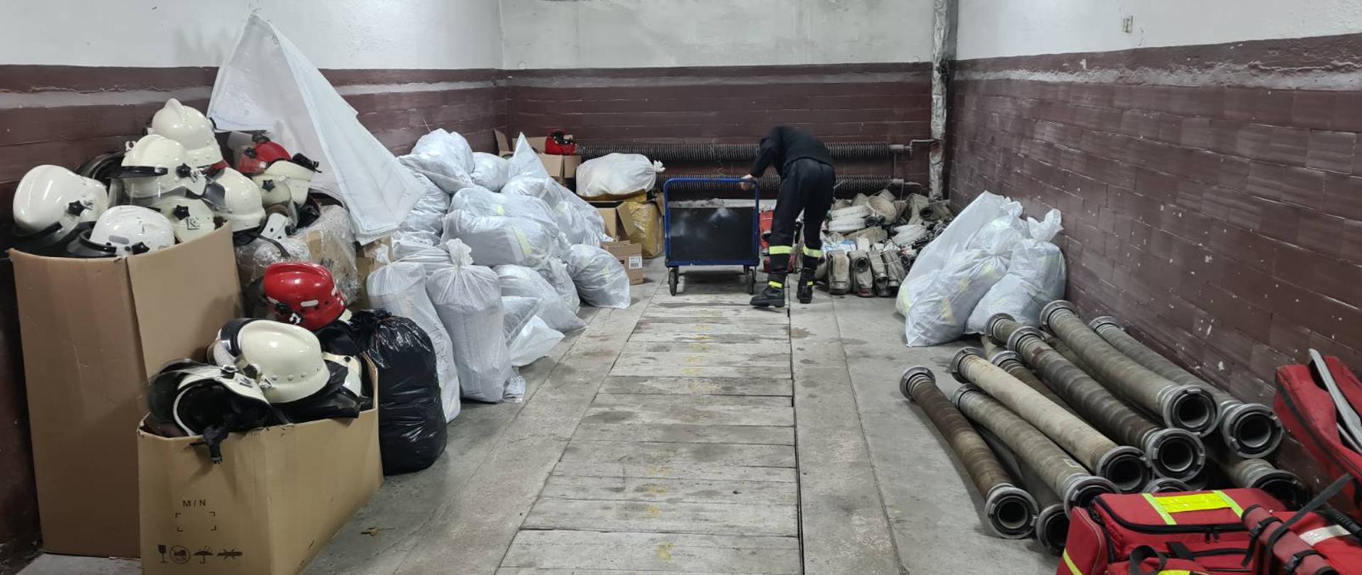 Zdjęcie przedstawia pomieszczenie garażowe, w którym zebrany po bokach jest sprzęt strażacki: białe i czerwone hełmy, węże, czerwone torby i inny zapakowany w białe worki. 