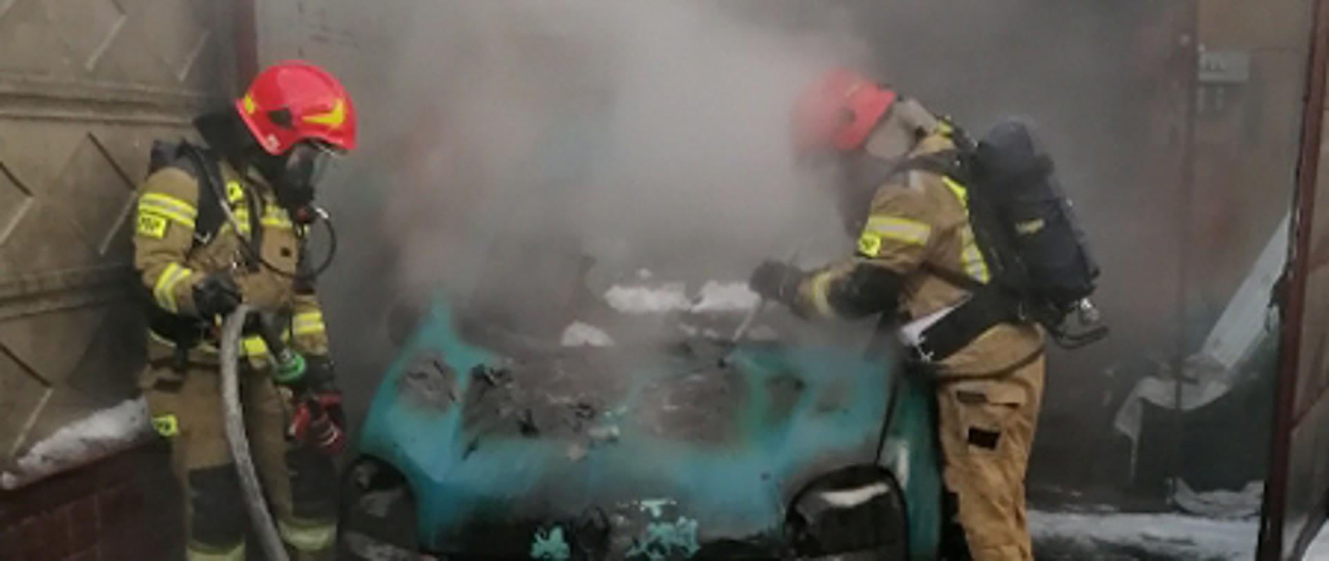 Pożar samochodu osobowego przed garażem. Po obu stronach pojazdu strażacy w aparatach powietrznych dogaszają samochód.