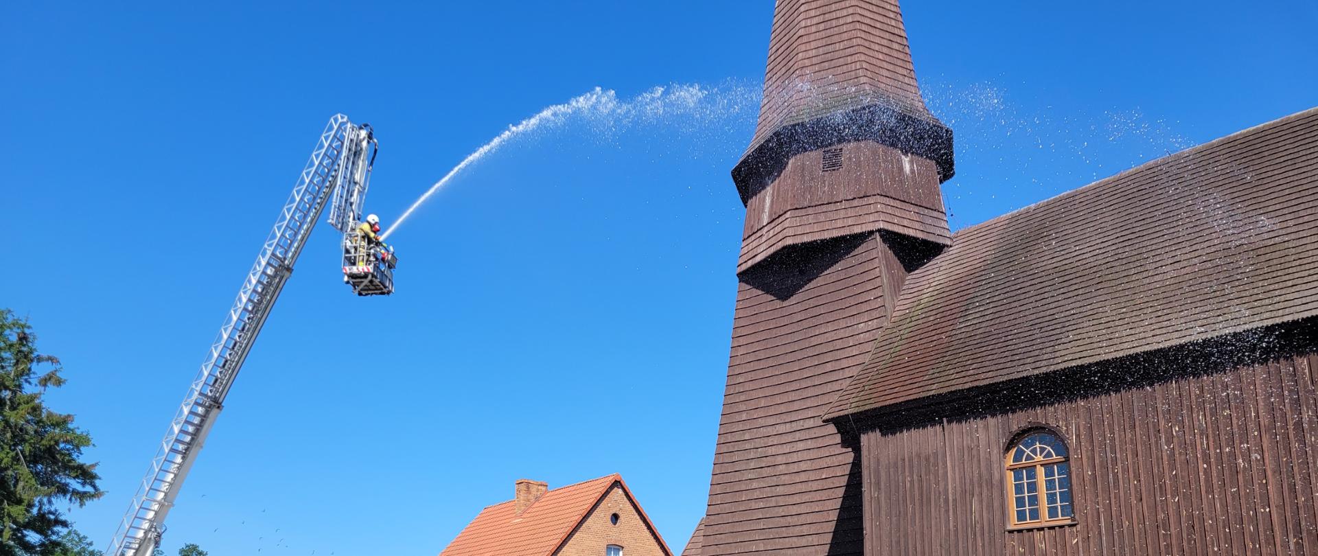 Słoneczna pogoda, podnośnik strażacki na betonowym podłożu, w oddali drewniany zabytkowy kościół. Z podnośnika podawana jest woda na dach kościoła na tle błękitnego nieba.