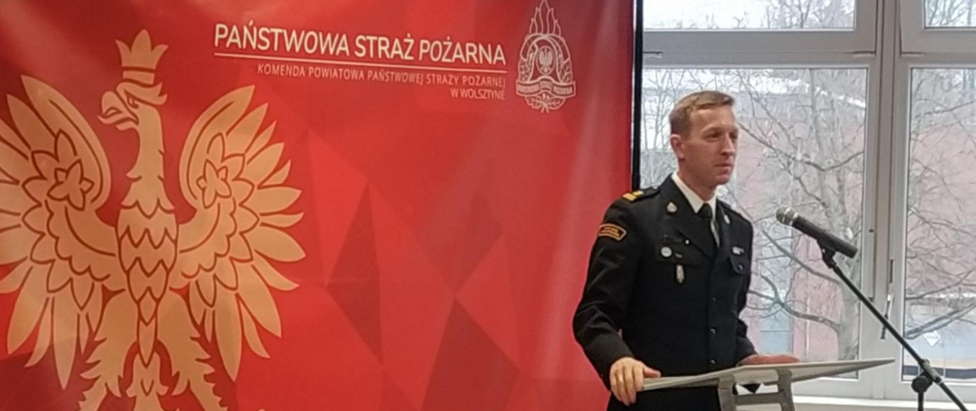 strażak sto przy mównucy, przemawia, za nim czerowna ścianka tekstylna z orlem i napisem państwowa straż pożarna