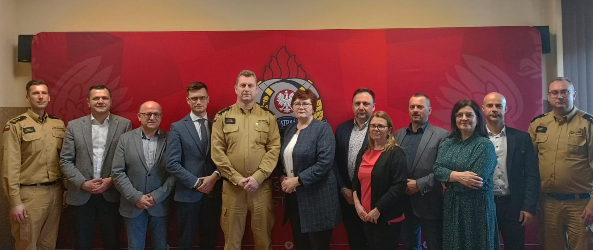 Zdjęcie grupowe uczestników spotkania Komisji Samorządności i Bezpieczeństwa Powiatu Częstochowskiego. W tle czerwona ścianka reklamowa Państwowej Straży Pożarnej.