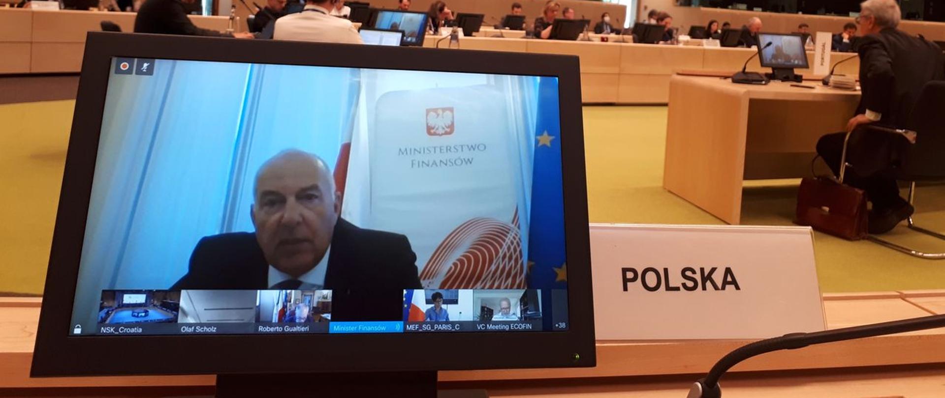 Minister Kościński na ekranie komputera podczas wideokonferencji, obok monitora napis Polska.