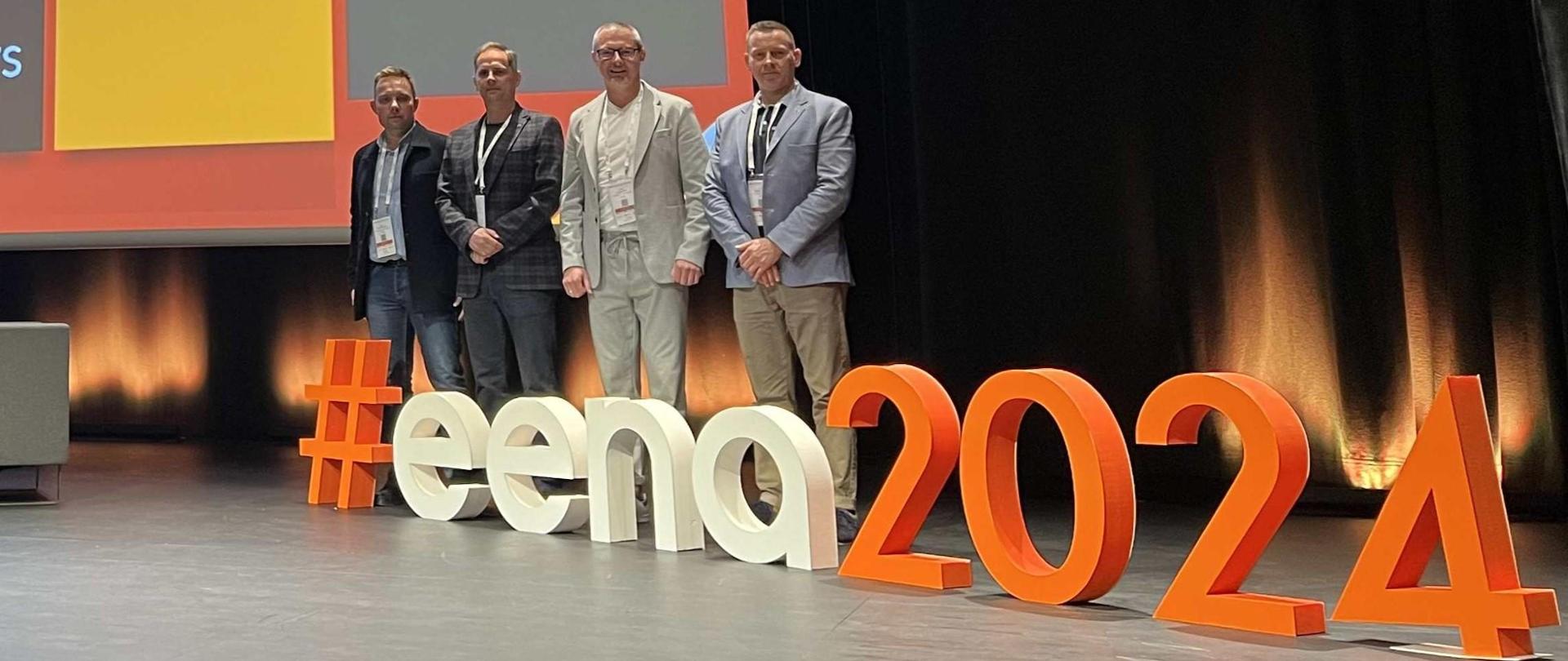 Przedstawiciele Państwowej Straży Pożarnej na Konferencji Europejskiego Stowarzyszenia Numeru Alarmowego 112. Czterech uczestników konferencji stoi na scenie a przed ich nogami skrót EENA oraz rok.