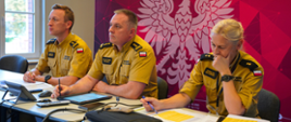 Panowie Komendanci i Naczelnik Wydziału Kadr uczestniczą w sali prezydialnej KW PSP w wideokonferencji, zajmując miejsca za stołem, przed telewizorem.