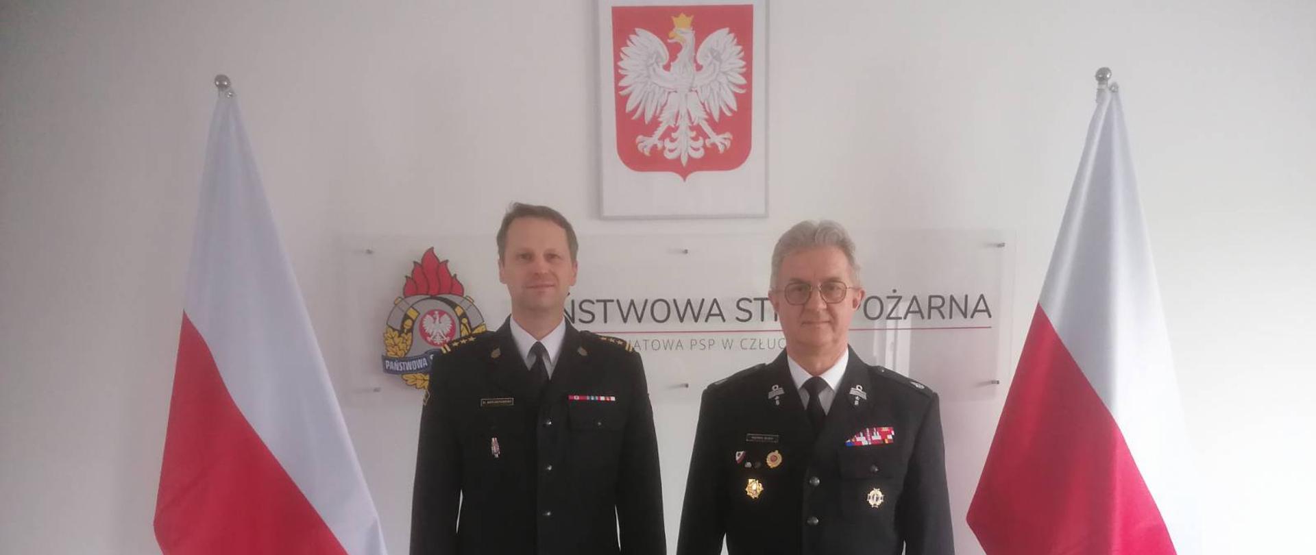 Komendant Powiatowy Państwowej Straży Pożarnej w Człuchowie st. bryg. Ernest Szczepański z dh. Markiem Buzą - Prezesem Zarządu Oddziału Wojewódzkiego Związku Ochotniczych Straży Pożarnych Rzeczypospolitej Polskiej województwa pomorskiego stojący między flagami państwowymi na tle logo komendy.
