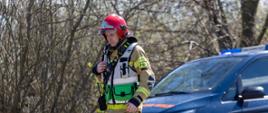 Strażak ratowniczy w czerwonym hełmie i specjalistycznym ubraniu ochronnym stoi przy drodze obok pojazdu służb ratunkowych. W tle widać bezlistne drzewa, a na pierwszym planie rozłożony sprzęt ratowniczy.