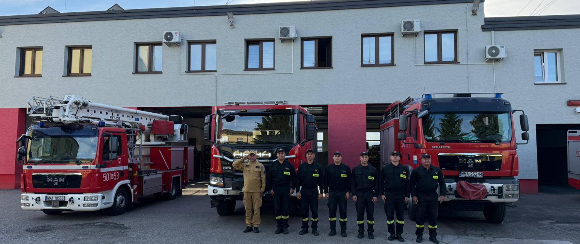 Strażacy z KP PSP w Makowie Mazowieckim oddają hołd zmarłemu druhowi OSP. z tyłu widać trzy wozy strażackie oraz budynek komendy.