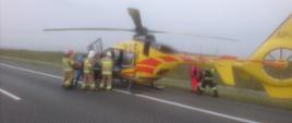 Na zdjęciu widoczny śmigłowiec Lotniczego Pogotowia Ratunkowego - ustawiony czołem maszyny przeciwnie do fotografującego - koloru żółtego (z czerwonymi pasami ciągnącymi się od drzwi do ogona), do którego strażacy pomagają włożyć osobę poszkodowaną na nosze. Pięciu strażaków wraz z obsadą LPR wkładają poszkodowanego do śmigłowca od jego lewej strony. Czterech strażaków w umundurowaniu specjalnym koloru piaskowego (dwóch strażaków w białych i dwóch w czerwonych hełmach). Za śmigłowcem widoczny członek załogi LPR na wysokości połowy ogona wraz z idącym za nim strażakiem w umundurowaniu specjalnym koloru czarnego.
Śmigłowiec marki Eurocopter na zdjęciu wylądował na pasie awaryjnym (asfalt) autostrady. W tle widoczny osiatkowany płot autostrady oraz pas zieleni.