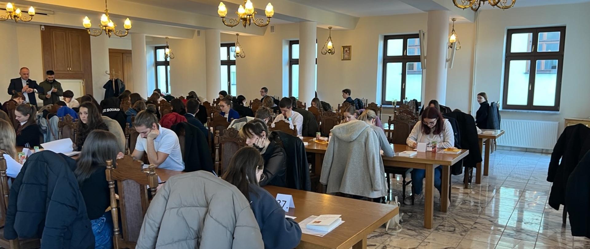 Zdjęcie przedstawia grupę młodzieży rozwiązującej zadania na olimpiadzie teologicznej.