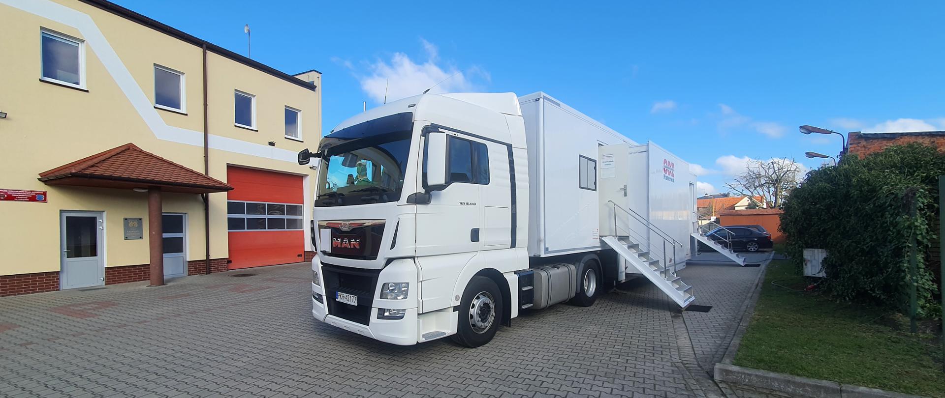 Na placu komendy powiatowej rozstawiony został mobilny symulator jazdy z ośrodka szkolenia jazdy, Na tle symulatora znajdującego się na podwoziu ciężarowym widoczny budynek komendy powiatowej. 