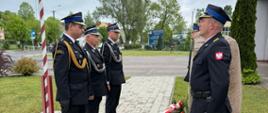 Trzech strażaków w pełnym umundurowaniu oddaje hołd poległym druhom; w tle widoczny jest plac komendy otoczony drzewami