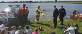 Strażacy demonstrują dzieciom zebranym przy zbiorniku wodnym metody ratowania osób tonących z brzegu. Obok samochód strażacki oraz łódź motorowa.