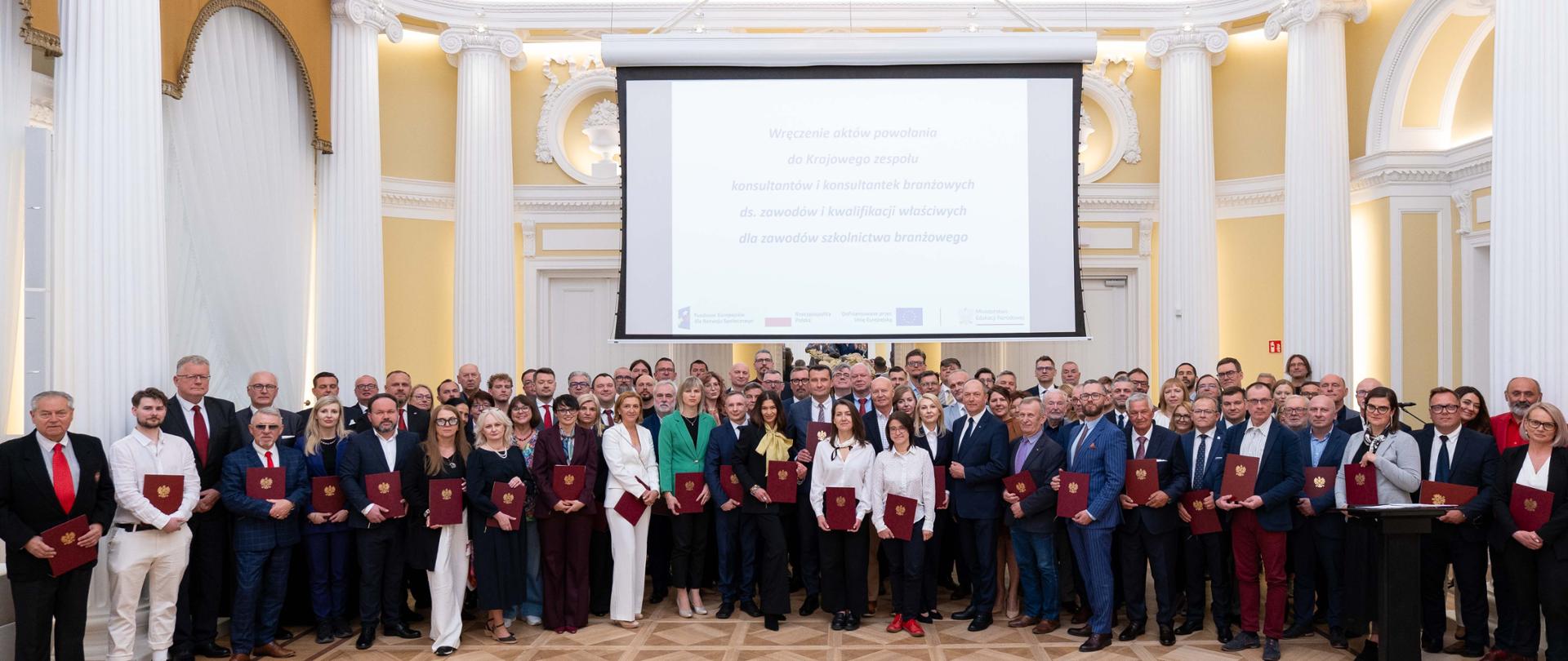 Liczna grupa osób pozuje do wspólnego zdjęcia w eleganckiej, jasnej sali z klasycznymi kolumnami. Uczestnicy stoją w rzędzie, wielu z nich trzyma w dłoniach bordowe teczki z aktami powołań. Nad grupą wisi duży ekran projekcyjny z wyświetlonym tekstem informującym o wręczeniu aktów powołania do Krajowego Zespołu Konsultantów Branżowych.