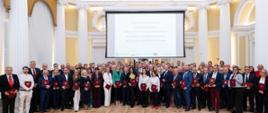 Liczna grupa osób pozuje do wspólnego zdjęcia w eleganckiej, jasnej sali z klasycznymi kolumnami. Uczestnicy stoją w rzędzie, wielu z nich trzyma w dłoniach bordowe teczki z aktami powołań. Nad grupą wisi duży ekran projekcyjny z wyświetlonym tekstem informującym o wręczeniu aktów powołania do Krajowego Zespołu Konsultantów Branżowych.
