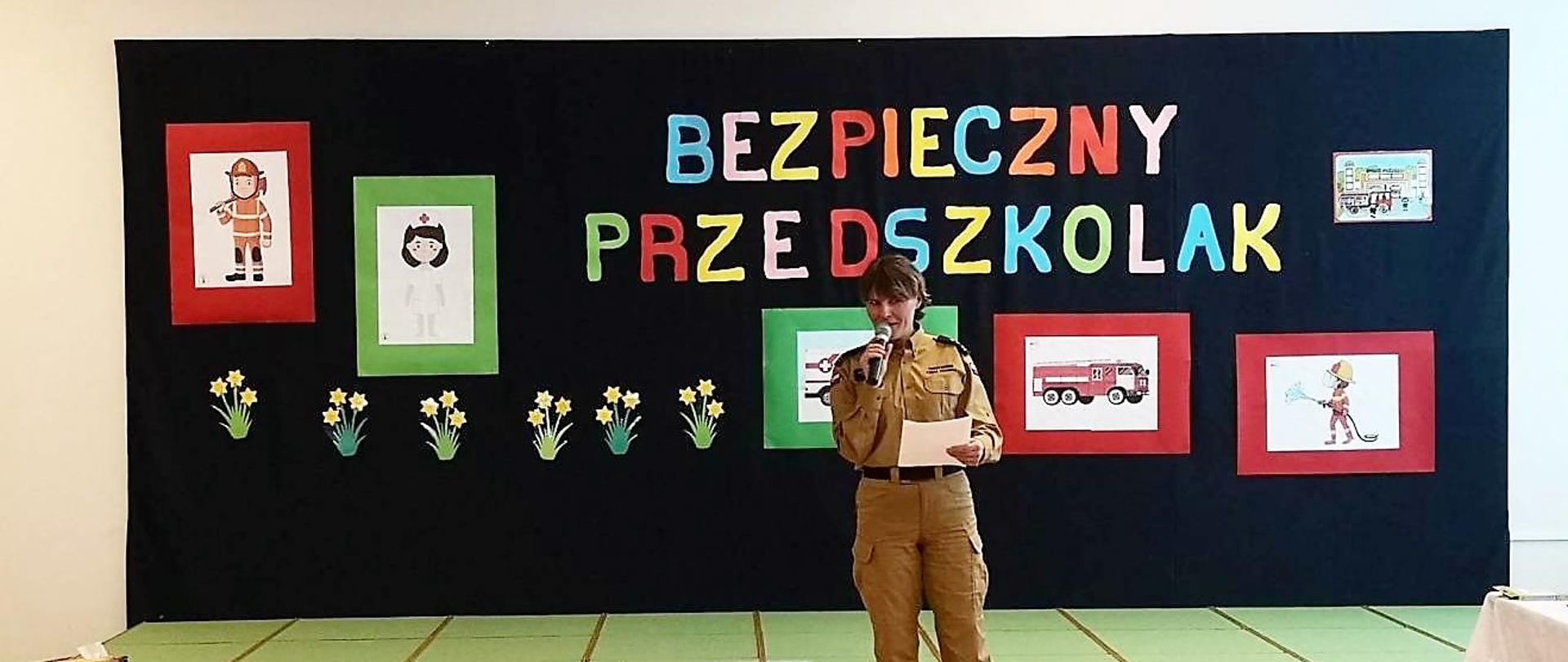 Pani strażak ubrana w mundur służbowy w kolorze musztardowym trzyma w dłoni mikrofon, przez który przemawia do zgromadzonych dzieci. Za nią znajduje się czarna ścianka, na której znajduje się kolorowy napis "BEZPIECZNY PRZEDSZKOLAK" a dokoła napisu przyczepione są rysunki dzieci o tematyce strażackiej.