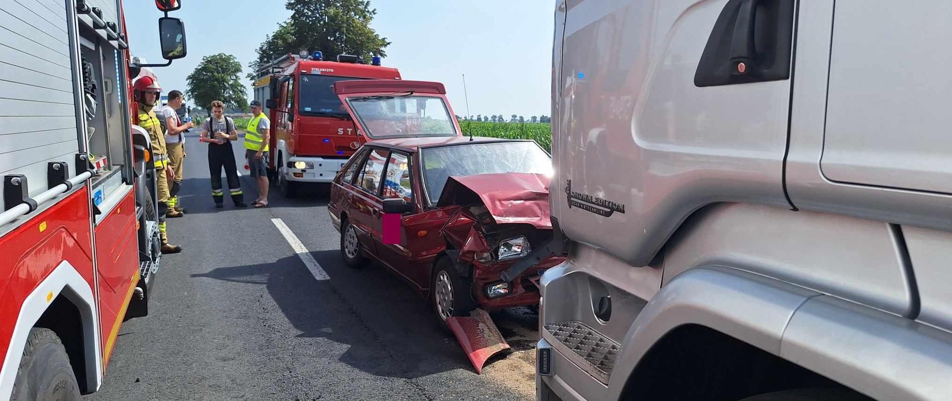 Na jednym z pasów ruchu drogi krajowej, na przeciwko siebie stoją samochód osobowy w kolorze wiśniowym oraz srebrny ciągnik siodłowy. Przód auta osobowego jest znacznie uszkodzony. Za nim stoi jeden wóz strażacki, drugi znajduje się na pasie obok. Pomiędzy samochodami widoczni są strażacy uczestniczący w akcji. Widoczna jest jego przednia lewa strona, w znacznym stropniu uszkodzona.Wokół samochodu rozsypany jest sorbent.