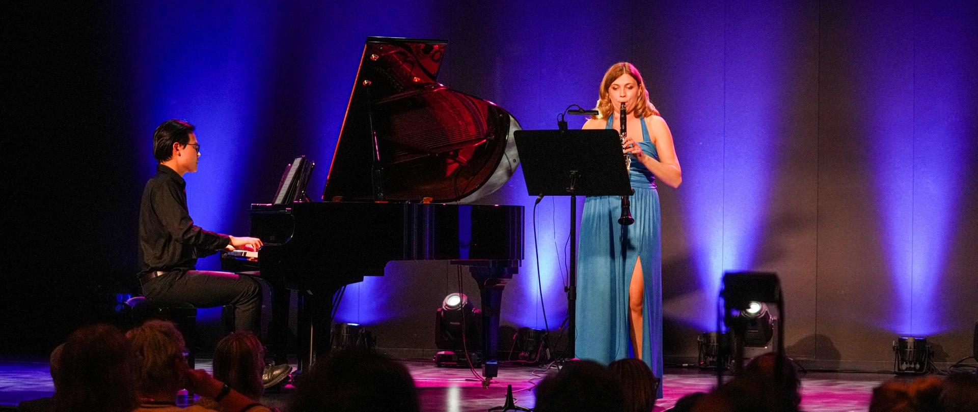Klarnecistka w błękitnej sukni gra na klarnecie przy akompaniamencie pianisty na oświetlonej fioletowym światłem scenie przed siedzącą publicznością.