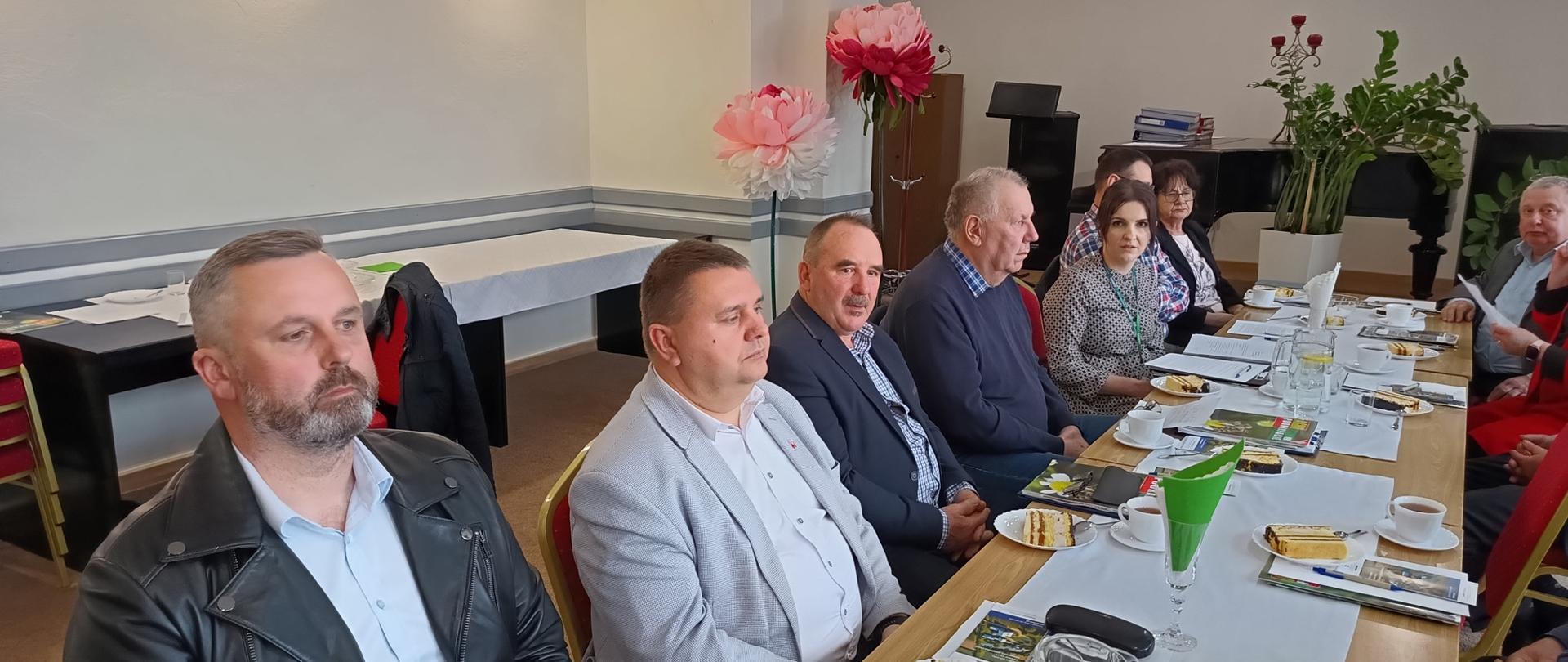 Grupa kilkunastu dorosłych osób siedzi przy długim, prostokątnym stole w jasnej sali spotkań. Uczestnicy, głównie w średnim i starszym wieku, ubrani są w eleganckie lub półformalne stroje – marynarki, koszule i swetry. Na stole znajdują się filiżanki z kawą i herbatą, talerzyki z kawałkami ciasta, szklanki z wodą oraz dokumenty i notatniki. Jedna z osób na pierwszym planie zapisuje coś w zeszycie. W tle widoczne są rośliny doniczkowe, fortepian oraz dekoracyjne kwiaty z papieru. Ściany są jasne, z zamontowanym oświetleniem punktowym i widocznymi rurami wentylacyjnymi przy suficie. Atmosfera sugeruje formalne spotkanie lub naradę.