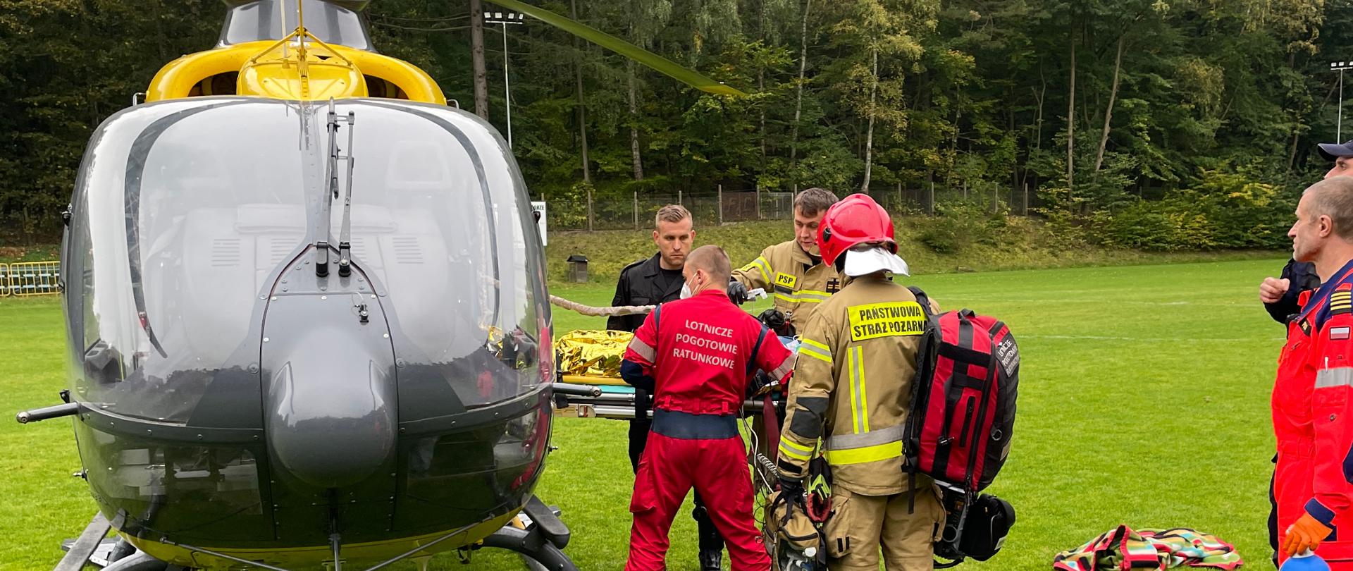 Śmigłowiec Hemes lotniczego pogotowia ratunkowego stojący na płycie boiska. DO śmigłowca strażacy i ratownicy wkładają poszkodowaną osobę na noszach