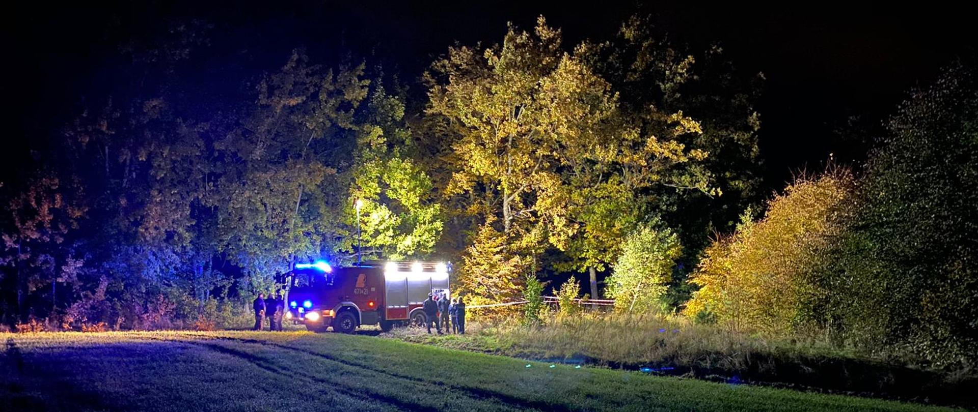 Średni samochód ratowniczo-gaśniczy w oddali w lesie. Przy samochodzie Państwowej Straży Pożarnej widoczni funkcjonariusze Policji prowadzący działania.
