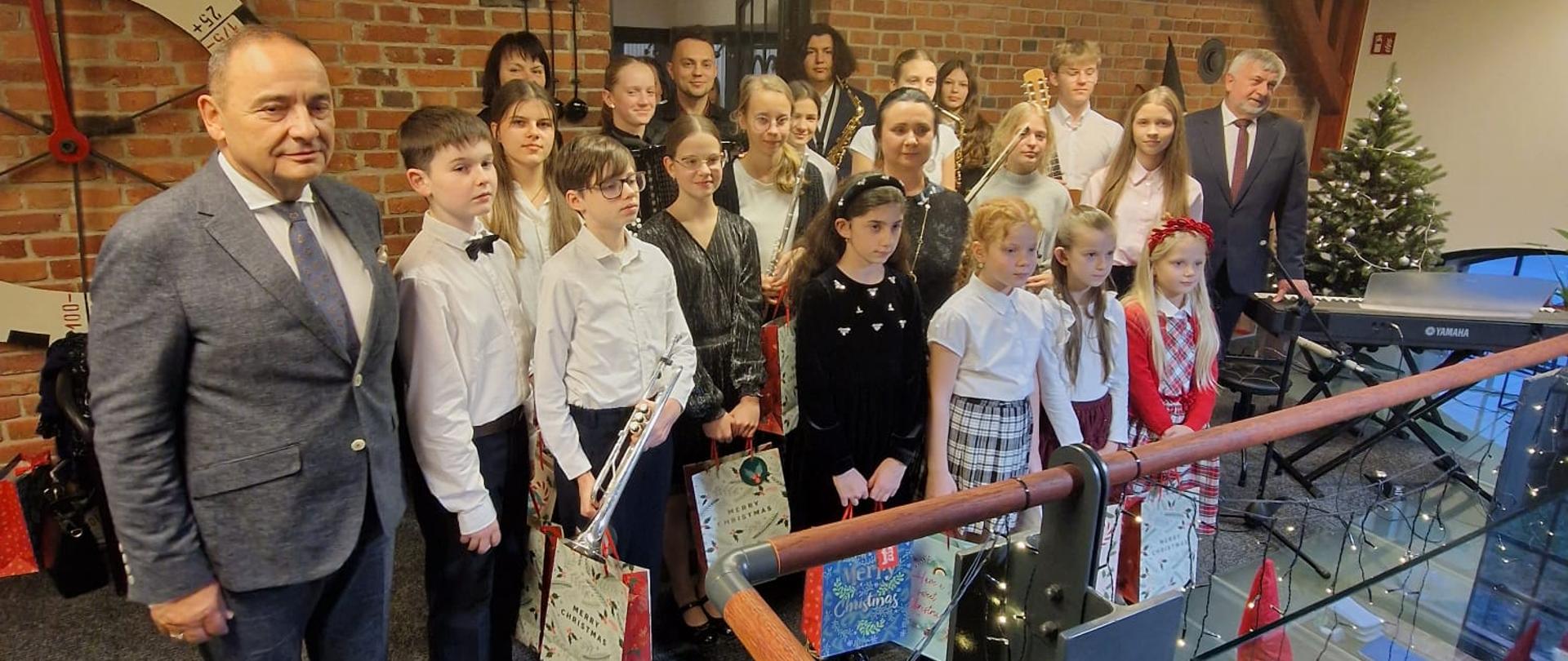 Zdjęcie grupowe wykonane w przestronnym wnętrzu z czerwonej cegły. Na pierwszym planie stoi grupa dzieci ubranych odświętnie – chłopcy w białych koszulach, niektóre dziewczynki w błyszczących sukienkach, inne w strojach wizytowych. Część z nich trzyma świąteczne torby z upominkami oraz instrumenty muzyczne. Wśród dzieci widoczne są również dwie dorosłe osoby w garniturach. Tło zdobią świąteczne akcenty: mała choinka, girlandy lampek i figurka świątecznego skrzata. 