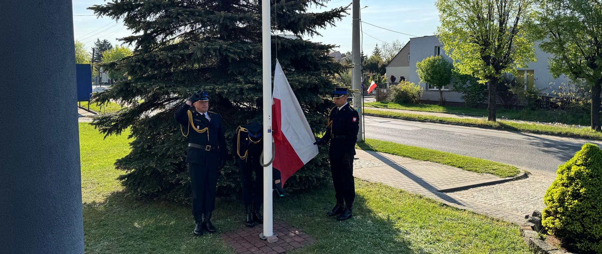 Na zdjęciu widoczni są trzej strażacy w umundurowaniu galowym stojących przy maszcie. Strażacy dokonują uroczystego podniesienia flagi państwowej na maszt