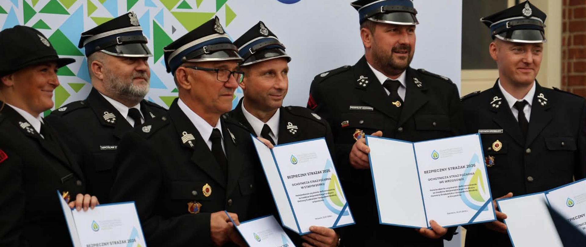 Grupa strażaków Państwowej Straży Pożarnej w galowych mundurach pozuje do wspólnego zdjęcia, trzymając otwarte teczki z certyfikatami programu „Bezpieczny Strażak 2026”. W tle znajduje się ścianka Wojewódzkiego Funduszu Ochrony Środowiska i Gospodarki Wodnej w Szczecinie.