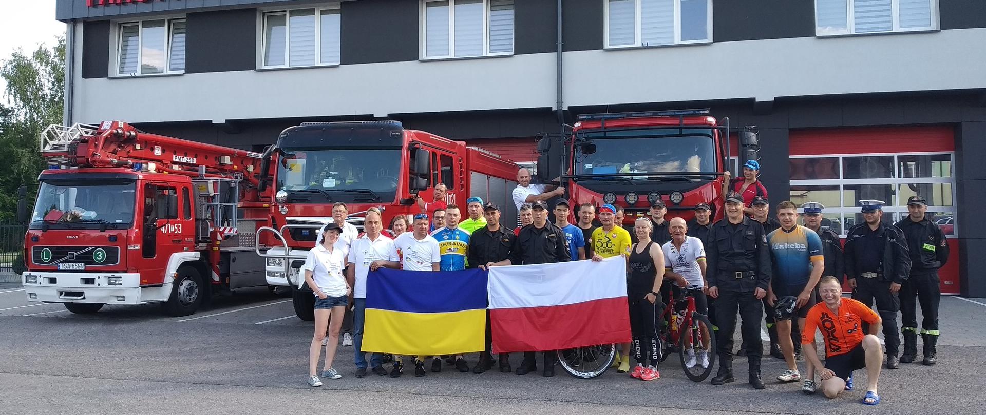 Zdjęcie zrobione na tle remizy i strażackich samochodów. Doz zdjęcia pozują strażacy i kolarze trzymający flagę Polski i Ukrainy