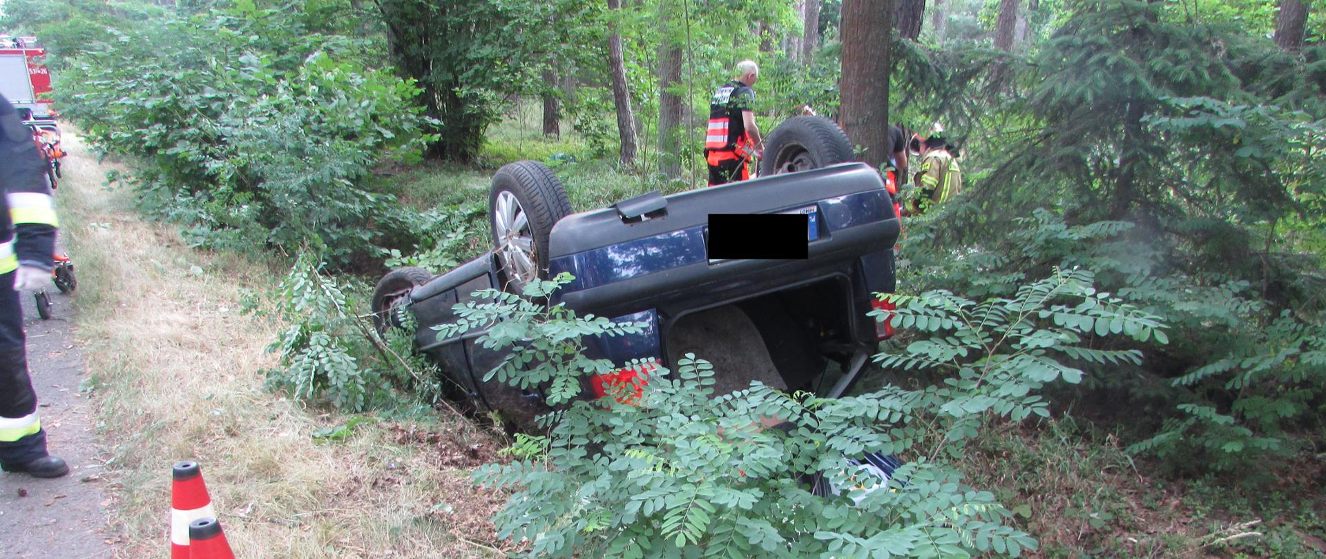 W rowie. w otoczeniu lasu, znajduje się pojazd po dachowaniu. W tle wśród drzew i krzewów znajdują się ratownicy medyczni i strażacy, którzy udzielają pierwszej pomocy poszkodowanemu. Z lewej strony widać dwa pachołki drogowe.