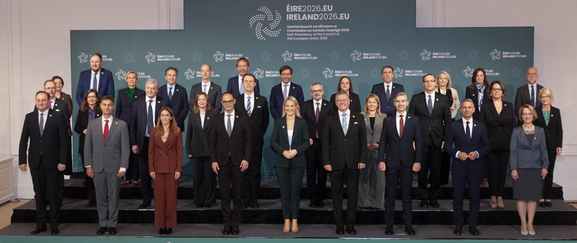 Zdjęcie wszystkich uczestników nieformalnego posiedzenia Rady UE ds. Zagranicznych w formacie ministrów ds. handlu. Delegaci stoją w trzech rzędach, w tle ścianka z logotypami prezydencji irlandzkiej