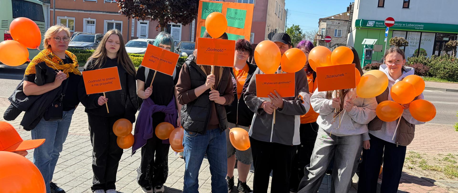 Grupa młodzieży i dorosłych stoi na chodniku podczas marszu społecznego w słoneczny dzień. Uczestnicy trzymają pomarańczowe balony i tabliczki z hasłami dotyczącymi godności i szacunku. W tle widać kolorowe kamienice oraz ulicę miasta.