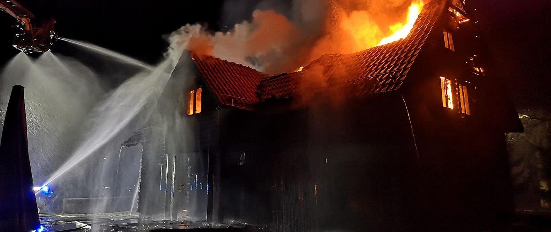 Płonący drewniany budynek widziany od frontu. Z dachy i z okien wydostają się płomienie. Z lewej strony widoczny kosz podnośnika hydraulicznego z którego strażacy podają wodę na płonący dach budynku.